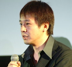 濃い緑色のスーツジャケットを着て、茶色の髪をした日本人男性がマイクに向かって話している肖像画。