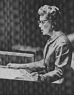 スーツジャケットを着て眼鏡をかけた短髪の女性が演壇で書類を読んでいるモノクロ写真