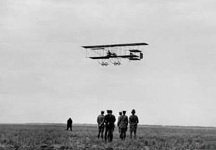 複葉機が畑の上を低空飛行し、数人の男たちがそれを見守っていた。