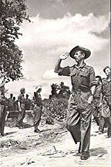 軍服を着てスラウチハットをかぶった兵士が数人の兵士を率いて行進している。彼は右側に静止している集団に敬礼をしている。