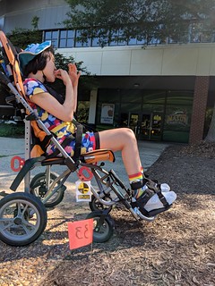 茶色の短い髪の少女が、ネオンオレンジ色のベビーカーに座っている。彼女は建物の前のコンクリートの歩道に立っている。