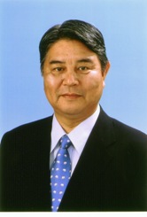 黒いスーツと白い水玉模様の青いネクタイを着用した桜井郁三の画像。背後には青い背景があります。
