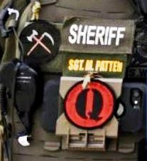 ある兵士の制服の詳細。赤い背景に黒い「Q」の文字が入ったワッペンと、斧と鎌が交差した黒い地に描かれた別のワッペンが見える。