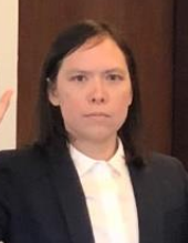 ツーピースのスーツを着てネクタイをしていないアジア系アメリカ人女性が右手を挙げている。