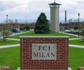 手前の赤レンガの看板には「FCI MILAN」と書かれています。後ろには、草の茂った丘の上に一本の緑の木が見えます。その右側には塔があります。