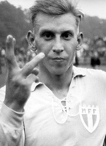 金髪の男性が、3本の指を立てて右手をカメラに向けています。彼は明るい色のサッカーユニフォームを着ています。