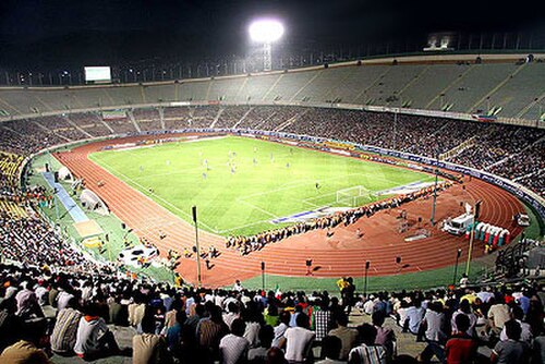 夜間のオープンルーフのスポーツアリーナ。芝生のフィールド(中央)と陸上トラック(フィールドの周囲)があり、座席のあるパビリオンにはサッカーの試合を観戦する観客がいっぱいです。