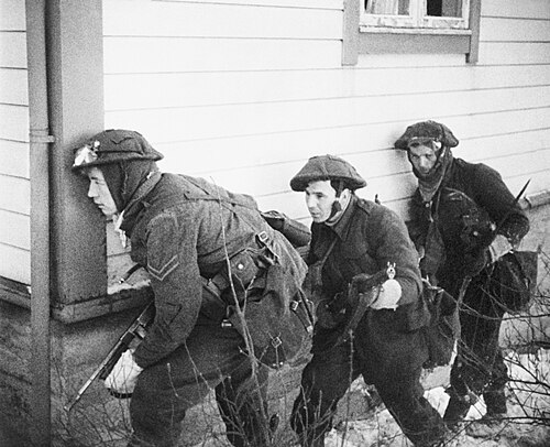 three British soldiers take cover at the corner of a house