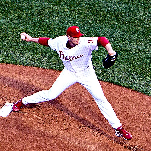 白い野球ユニフォームと赤い野球帽をかぶった男性が右手で野球ボールを投げている。ユニフォームの前面には青い縁取りの黒いブロック体文字で「トロント」と「34」と書かれており、左手には黒い野球グローブをはめている。