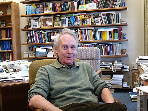 プルオーバーを着て、リラックスして、たくさんの本や書類のある書斎に座っているアントニー・ジェイムソンのカラー写真