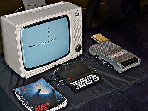 ZX81 は、「ZX81 BASIC プログラミング」マニュアルとカセット テープ レコーダーの間にあり、背景には白黒の Ferguson テレビが置かれています。