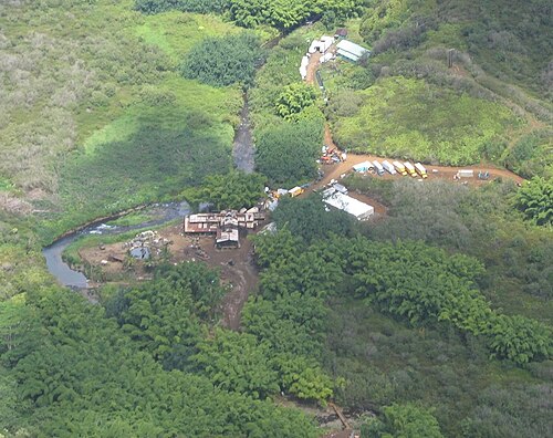 密林の中にある一群の建物を上から見た写真。建物の左側には川が流れ、建物の右側には移動中のトラックが見えます。建物から他の建物へは未舗装の道路が続いています。