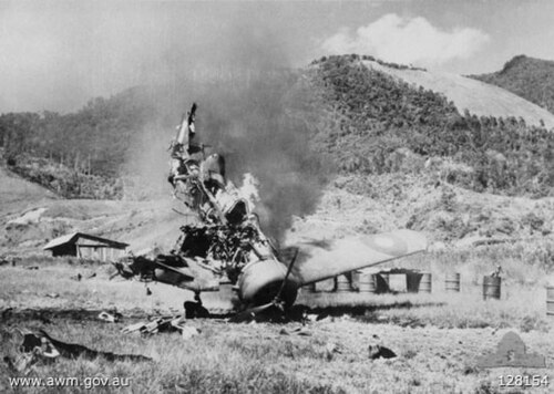 プロペラ機が機首を下に向けて炎上している。背景には44ガロン(約170リットル)のドラム缶が写っている。