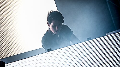 ポーター・ロビンソンは明るく照らされたスクリーンのあるテーブルの前に立っている。彼の後ろにはもう一つのスクリーンがある。