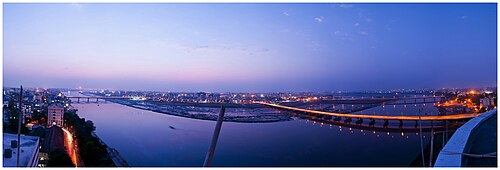 夕暮れ時のスラト市を流れるタピ川のパノラマ写真。部分的に日が差しており、街灯が灯っている。