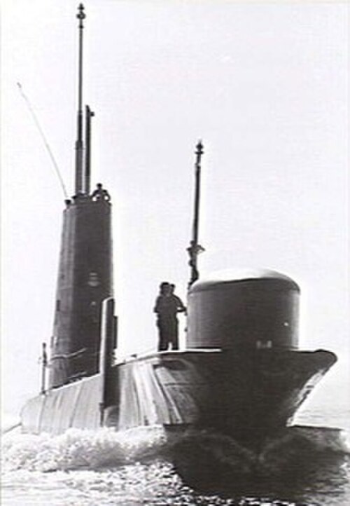 水面を航行中の潜水艦。船首のソナードームの近くに男性が立っており、帆の上には他の乗組員が見える。