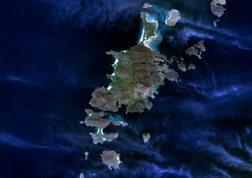 上から見た大きな濃い緑色の島は、濃い青色の水域にある小さな島々に囲まれています。薄い雲が部分的に視界を遮っています。