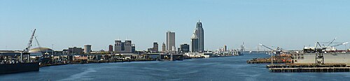 都市のスカイラインのパノラマビュー。手前には大きな川と港湾施設があります。遠くには、さまざまな高さの高層ビルが並ぶ都市のスカイラインが見えます。
