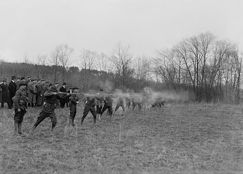 開けた野原に大勢の男性が立ち、実弾射撃訓練中にライフルを発砲している。射撃者の周囲には煙が見える。