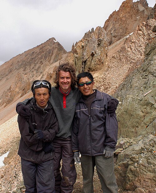 3人が外にカメラに向かって立っており、背景には山頂があり、真ん中の最も背の高い髭を生やした西洋人男性が、より背の低い2人のアジア人男性の肩に腕を回している。