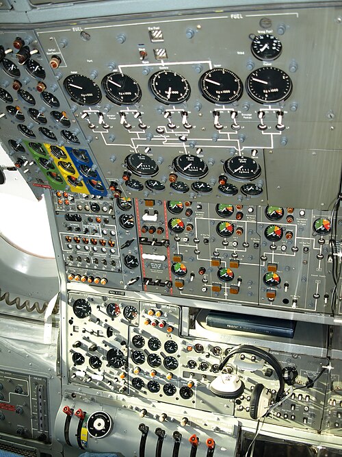 An airliner flight deck control panel with many instruments and switches. A pair of headphones hang from the panel.