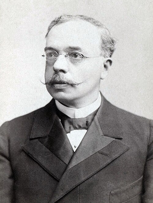 中年後半の男性の白黒画像。フォーマルな服装で小さな眼鏡をかけ、口ひげを生やしている。
