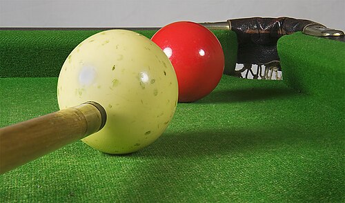 画像の右上隅のコーナーポケットの隣にある赤いスヌーカーボールの前方左側にある、穴だらけの白いボールのクローズアップ画像。画像の右下隅に、白いボールを打とうとしているキューの先端が見えています。