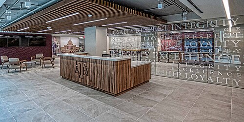 テキサス A&M のロゴが入った木製の机がコーヒー テーブルと椅子の横に置かれ、鏡張りの壁にはテキサス A&M の価値観が印刷されています。
