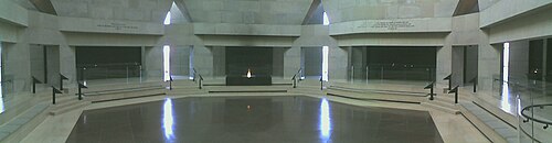 追悼ホールのパノラマビュー。赤いタイルの床、石灰岩の壁、黒いパネルのある六角形の部屋。前景の永遠の炎は、ヨーロッパの強制収容所からの灰が入った黒い箱に支えられています。