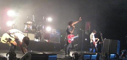 白い照明に照らされたステージには、ギタリスト2人、ドラマー1人、ベーシスト1人が立っている。ギタリストの1人がペダルを交換するために膝をついており、手前には複数のスピーカーが見える。