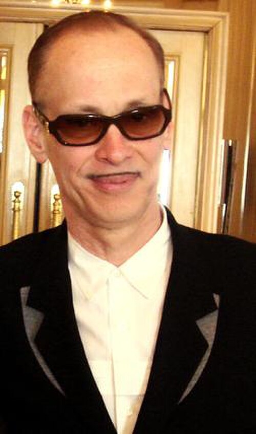 サングラスをかけ、黒いスーツを着て、小さな口ひげを生やした禿げ頭の男性。
