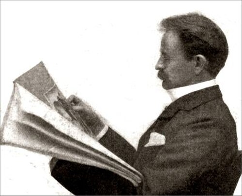 口ひげを生やした中年のビクトリア朝時代の男性が座って新聞を読んでいる。左から横顔を見たところ。