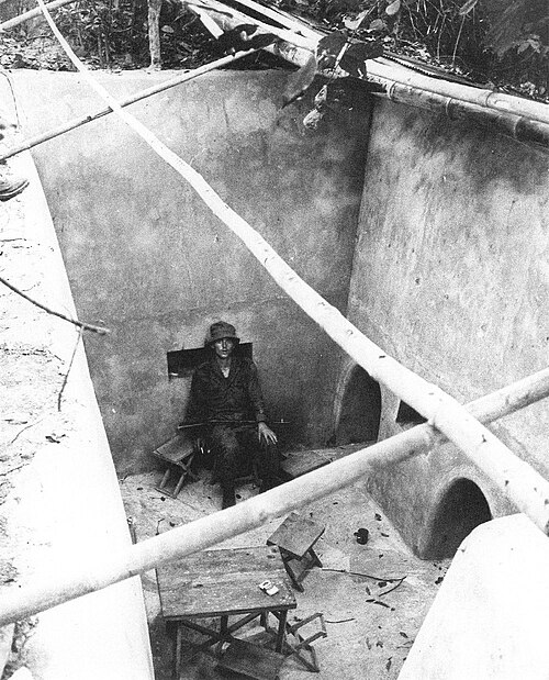 大きな露出した地下室で椅子に座っている兵士。右側の壁には 2 つの大きな穴があり、部屋には間に合わせのテーブルなど他の家具が散らかっている。