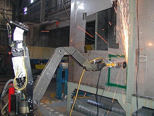 金属製のアームを備えたロボットが砥石を使って壁を削り、火花を上方に吹き出している。