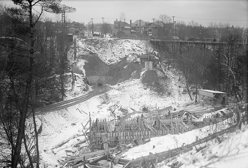 裸の谷を横切る工事。橋が建設中。地面は雪に覆われている。背景には、谷を越えて車を運ぶ鉄の架台がある。