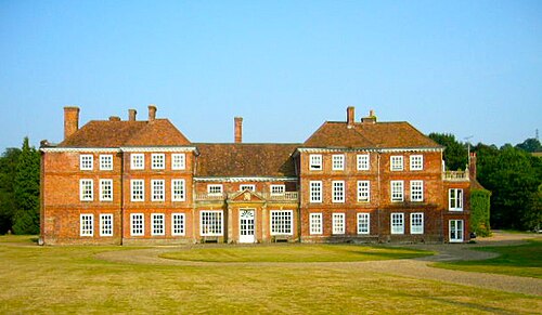 15 の上げ下げ窓がある大きな赤レンガの建物。中央の 3 つの間は 2.5 階建て、両側の外側の 6 つの間は 3.5 階建て、各側の最後の間はレンガで埋められています。刈り込まれた芝生の上に
