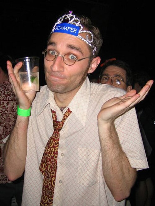 ティアラを着け、ペイズリー柄のネクタイを緩め、眼鏡をかけ、飲み物を持った男性がカメラに向かって肩をすくめている。