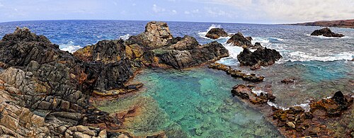 岩の多い海岸の写真。海からの澄んだ青緑色の水が自然のプールに満たされています。波がすぐそばで打ち寄せ、その向こうには青い海が広がっています。