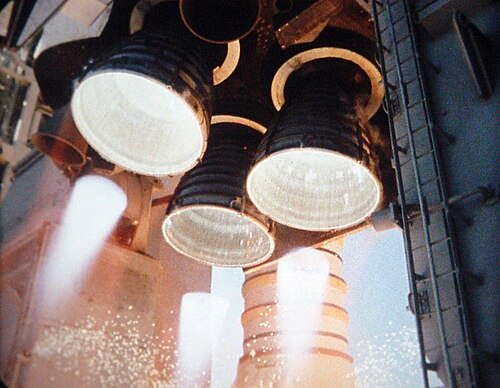 スペース シャトル オービターの後部構造から突き出ている 3 つのベル形のロケット エンジン ノズル。クラスターは三角形に配置されており、上部に 1 つのエンジン、下部に 2 つのエンジンがあり、上部エンジンの左右に 2 つの小さなノズルが見えます。3 つの大きなエンジンが点火しており、各ノズルから白熱した炎が噴出しています。背景には、スペース シャトルの左側の固体ロケット ブースター (白い円筒形のロケット) が見え、オービターの後部構造の左右には、2 つの大きな灰色の尾部サービス マストが見えます。