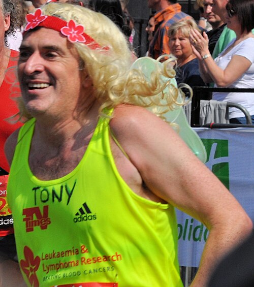 蛍光イエローのランニングトップを着て、金髪のかつらをかぶった男性のヘッドショット。