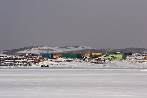 雪に覆われた平坦な平原に、建物群と遠くに小さな丘が見える。