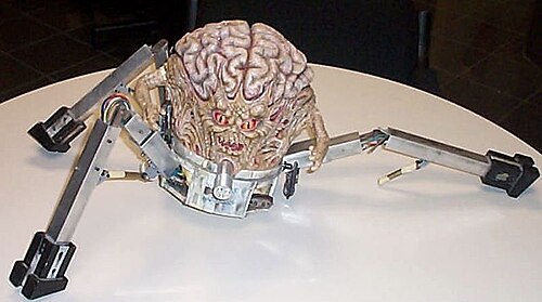 作動機構と露出した配線を備えた 3 本足のロボット ベースに搭載された脳のモデルの写真。脳には口と赤い目がある顔と、両側から伸びる小さな腕と、それをつかむ手があります。