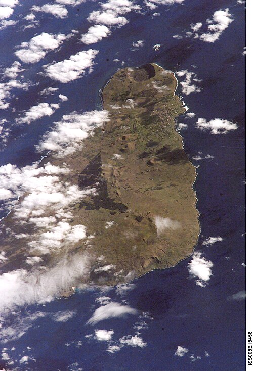 宇宙から撮影されたカラー写真。濃い青色の海に浮かぶ茶色がかった三角形の島が写っている。特に左側は白い雲に部分的に隠れている。