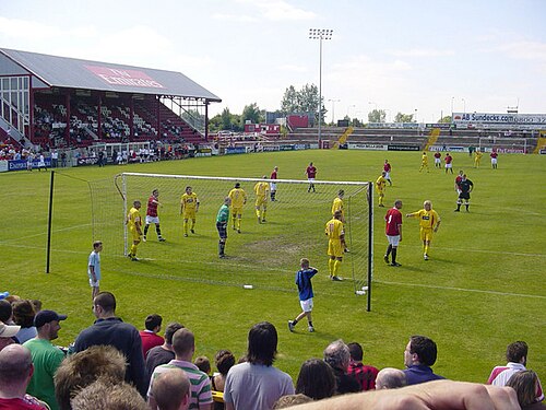 赤いユニフォームのFCユナイテッドと黄色いユニフォームのリー鉄道機械工学校の間でサッカーの試合が行われている。FCユナイテッドの観客が試合を観戦している。