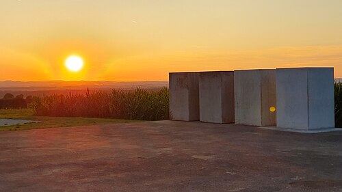 4つのコンクリートブロックがコンクリートの土台の上に立っており、周囲は起伏のある草地とトウモロコシ畑に囲まれている。
