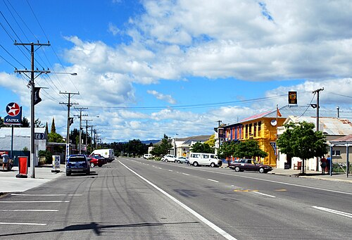 小さな町のまっすぐな道。両側にはお店、駐車中の車、そして時折小さな木が並んでいます。左側にはカルテックスのガソリンスタンド、右側には明るい青と黄色に塗られた 2 階建てのパブがあります。