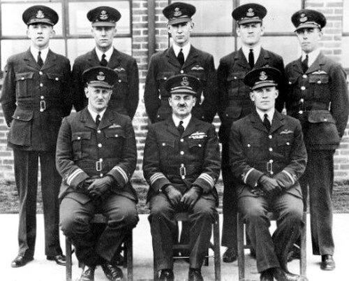 黒い軍服を着て山高帽をかぶった8人の男性の肖像画。5人が立っており、3人が座っている。