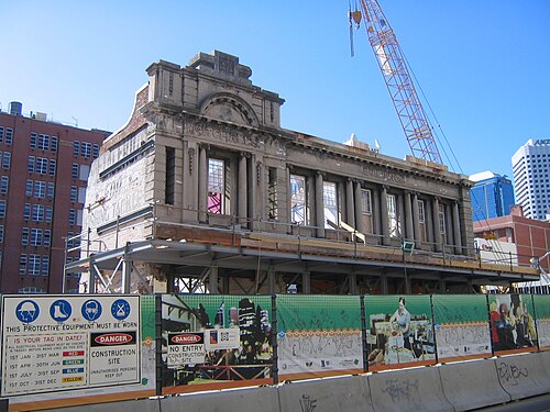 老朽化したように見える2階建ての建物で、周囲は工事現場に囲まれ、背後にはクレーンが立っている。