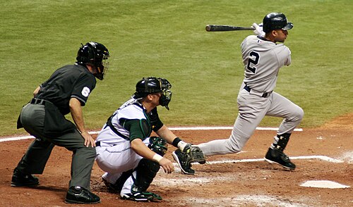 灰色のユニフォームを着た野球選手がフォロースルーでスイングし、その背後には白いユニフォームを着た相手チームのキャッチャーがしゃがみ、その背後には黒いユニフォームを着た審判が立っている。