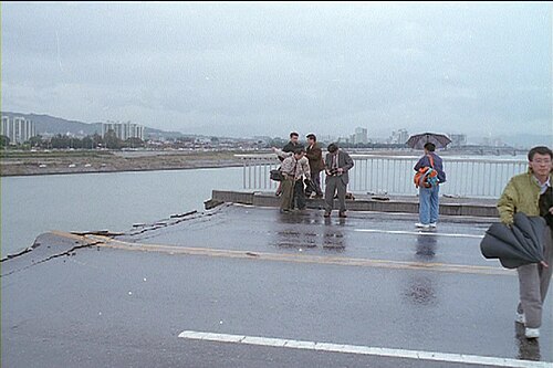 崩落後の橋の様子。傘をさした記者が数人見え、天気は曇り。道路は完全に途切れ、わずかに上向きに曲がっている。